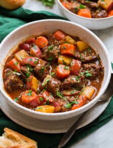 Beef Stew shown in a white serving bowl over a green cloth. Bread is shown to the side as a serving suggestion.