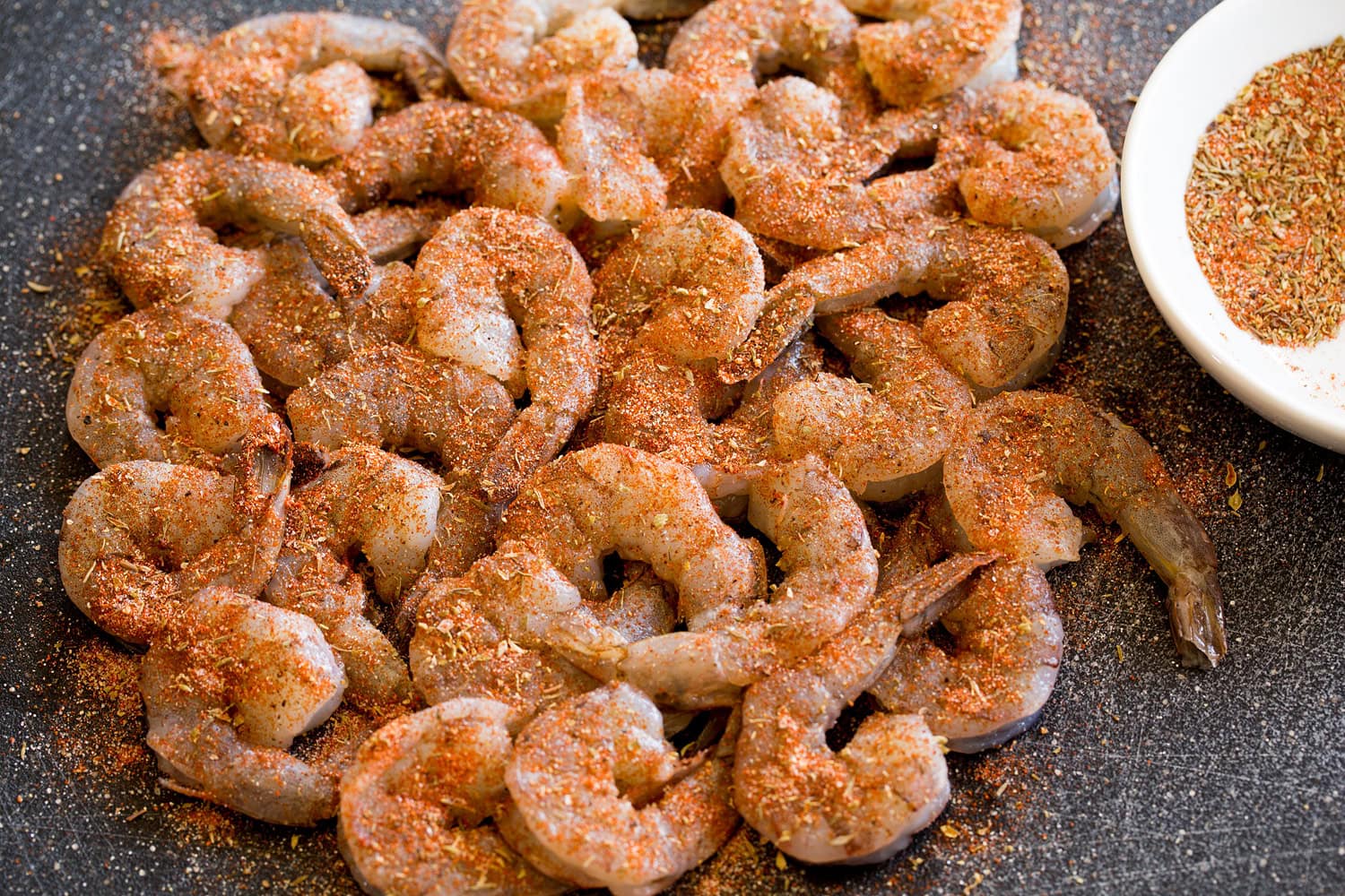 Raw shrimp sprinkled with blackening seasoning on a black cutting board.