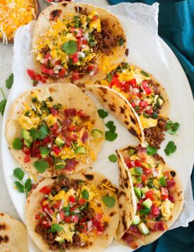 Breakfast tacos made with flour tortillas, chorizo, bacon, eggs, cheese, avocado and pico de gallo shown on a white oval platter from above.