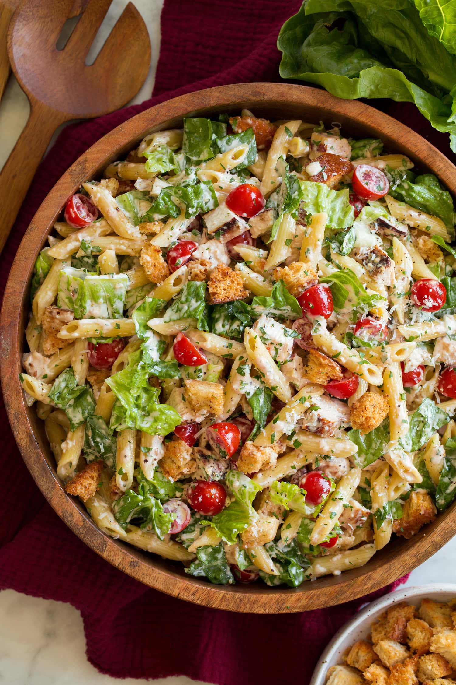 Grilled chicken caesar salad shown from overhead in a wooden bowl with romaine lettuce and wooden serving spoons to the side.