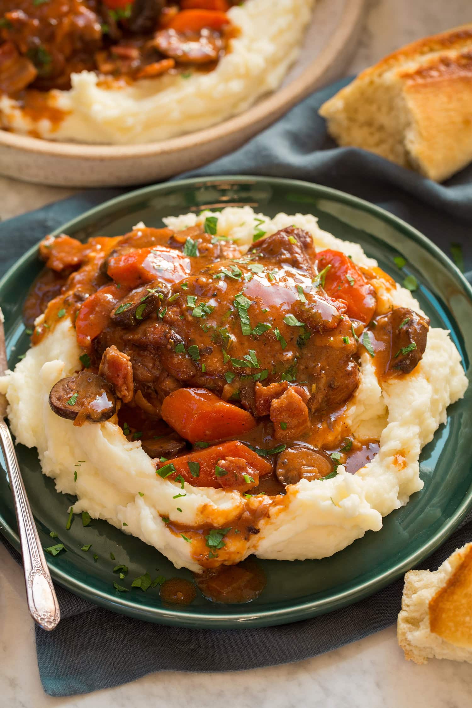 Coq au vin served over mashed potatoes on a green plate with bread to the side.