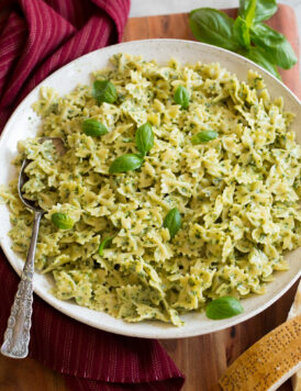 Creamy pesto pasta in a large white serving bowl with a red cloth and fresh basil to the side.