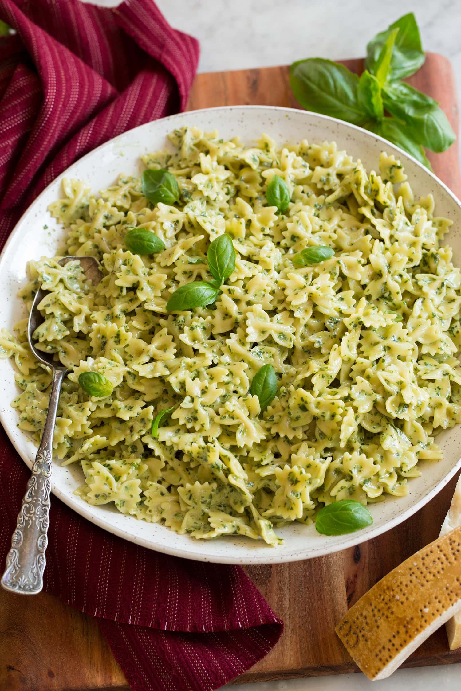 Creamy pesto pasta in a large white serving bowl with a red cloth and fresh basil to the side.