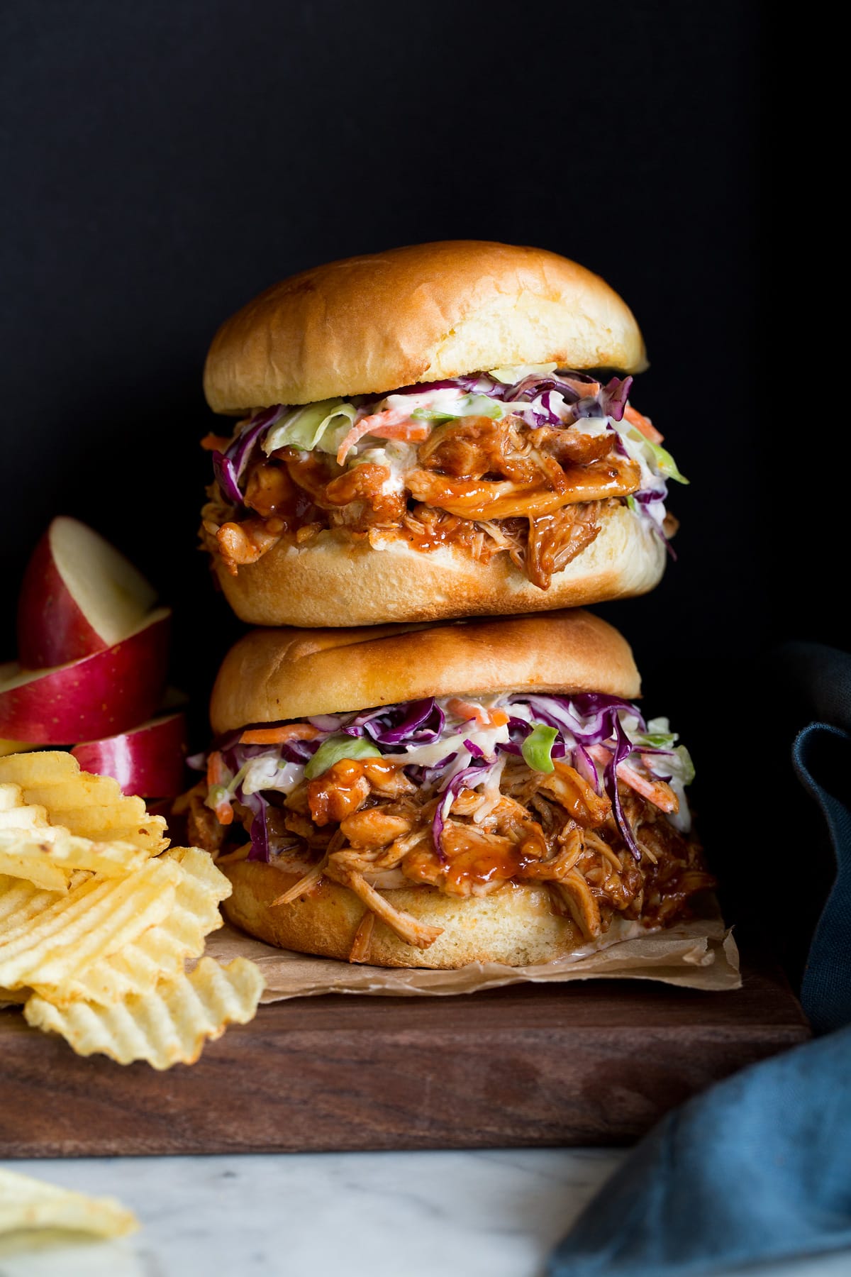 Photo: Two BBQ Pulled Chicken Sandwiches stacked on a wood platter. Apples slices and potato chips are shown to the side as serving suggestions.