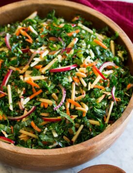 Photo: Kale salad with sliced apples, carrots, red onions, white cheddar and almonds shown in a wooden bowl over a marble surface. A red cloth is shown to the side and wooden salad spoons.