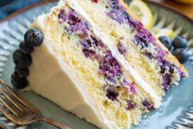 Slice of lemon blueberry cake with cream cheese frosting on a blue plate set over a wooden platter.