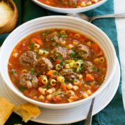 Serving of meatball soup in a white bowl over a white plate with bread to the side.