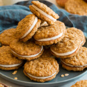 Stack of oatmeal cream pies on a blue plate with a blue cloth do the side and a glass of milk.