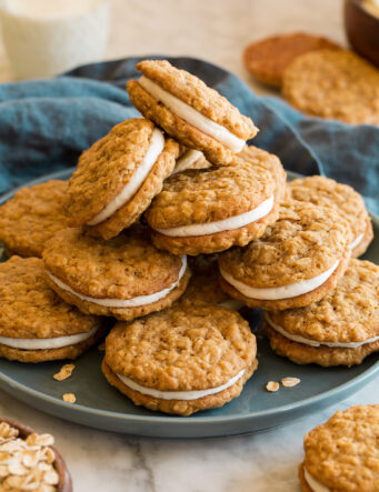 Stack of oatmeal cream pies on a blue plate with a blue cloth do the side and a glass of milk.