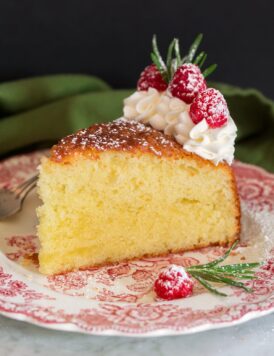 Photo of slice of olive oil cake decorated with whipped cream, raspberries and rosemary. Shown on a decorative red plate.