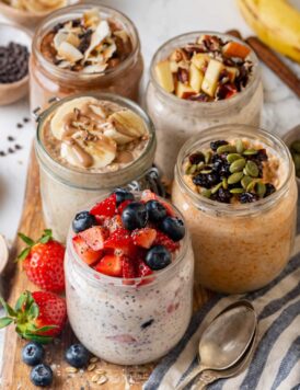 Overnight oats in mason jars, five different ways, on a wooden platter next to a spoon.