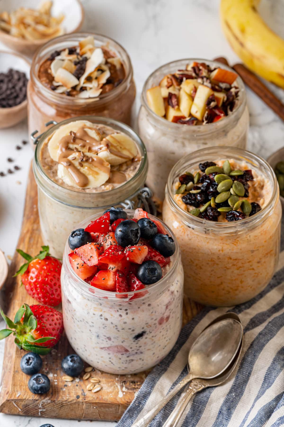 Overnight oats in mason jars, five different ways, on a wooden platter next to a spoon.