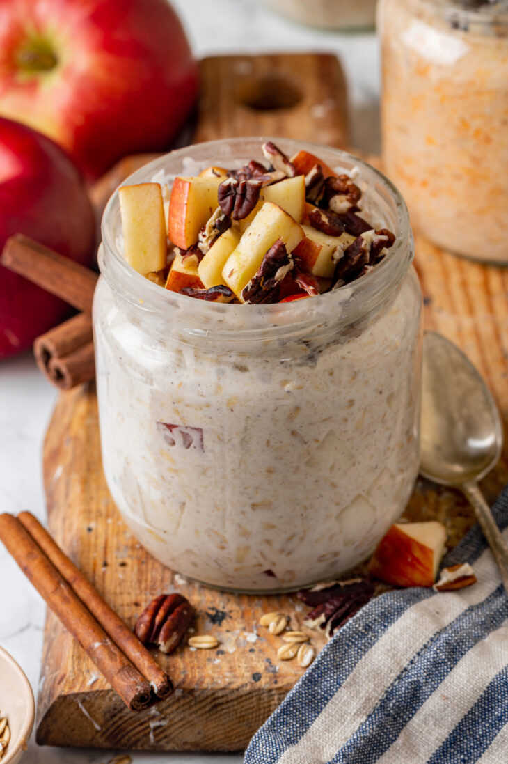 A mason jar of apple cinnamon overnight oats on a wooden platter, with an apple and cinnamon sticks in the background.