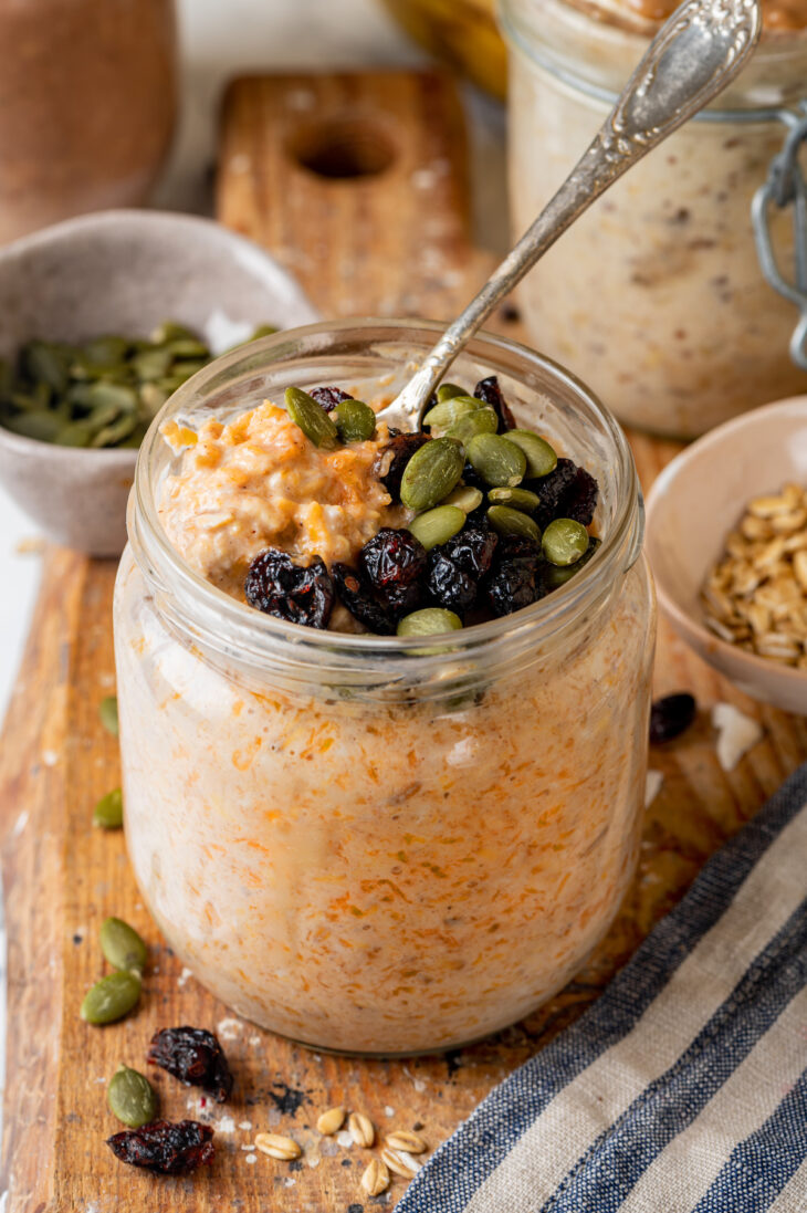 A spoonful of overnight oats held over a mason jar of pumpkin pie overnight oats on a wooden platter, with a small bowl of pepitas in the background.