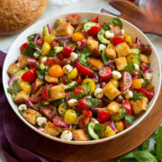 Panzanella salad in a white bowl over a wooden tray with a purple cloth and fresh basil and tomatoes shown to the sides.