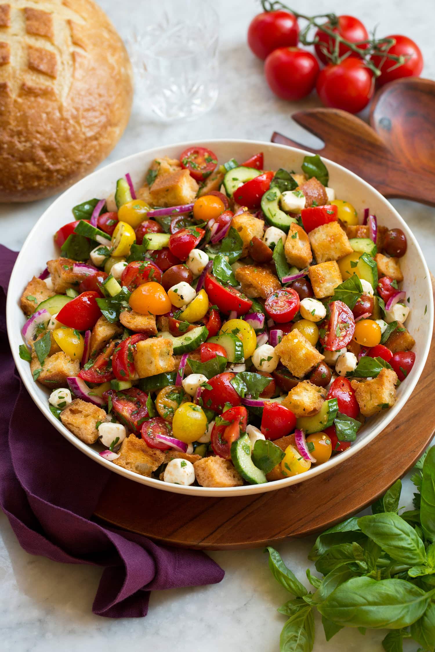 Panzanella salad in a white bowl over a wooden tray with a purple cloth and fresh basil and tomatoes shown to the sides.