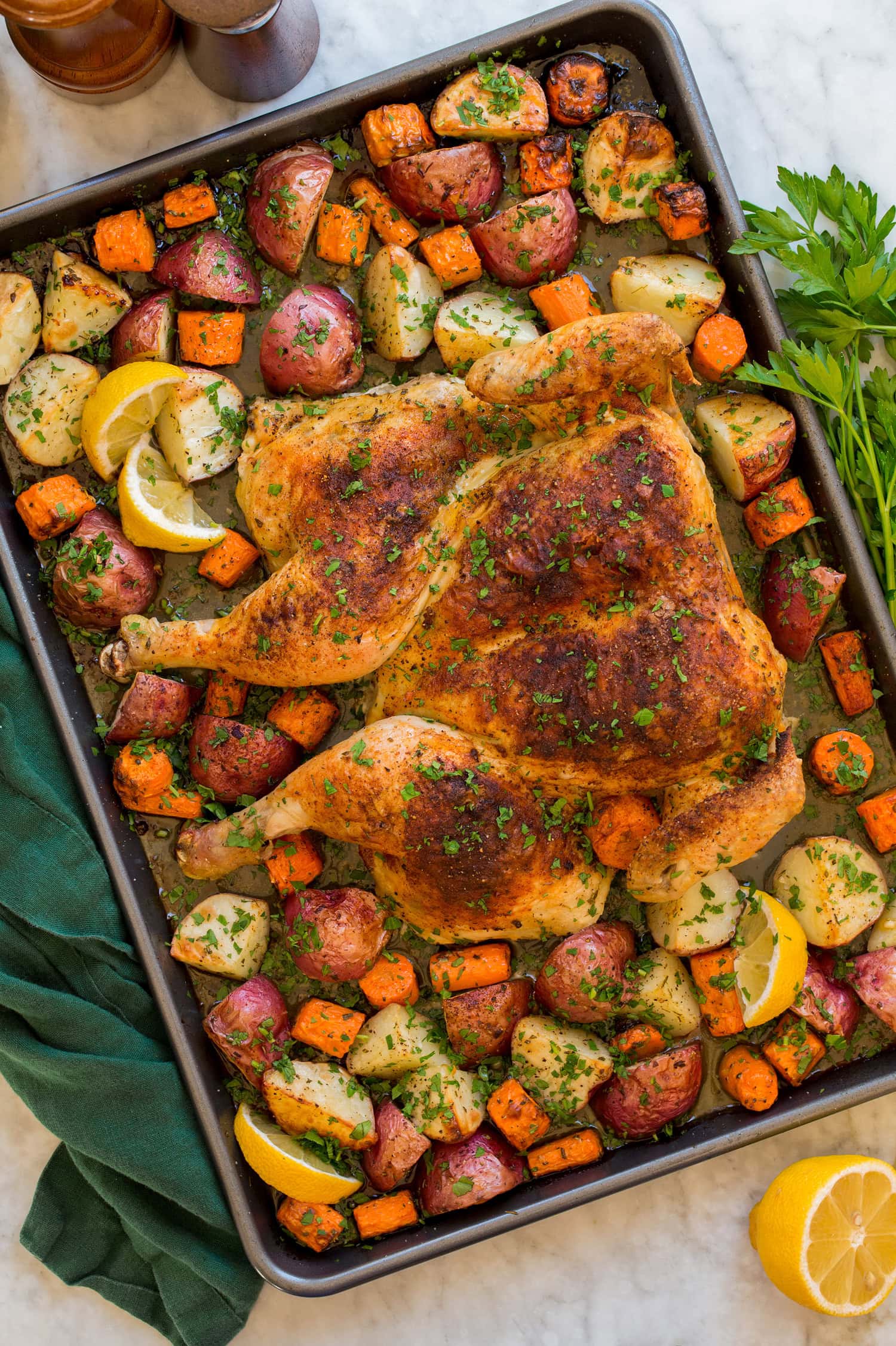 Spatchcock chicken with roasted vegetables on a dark rimmed baking sheet with a green cloth to the side.