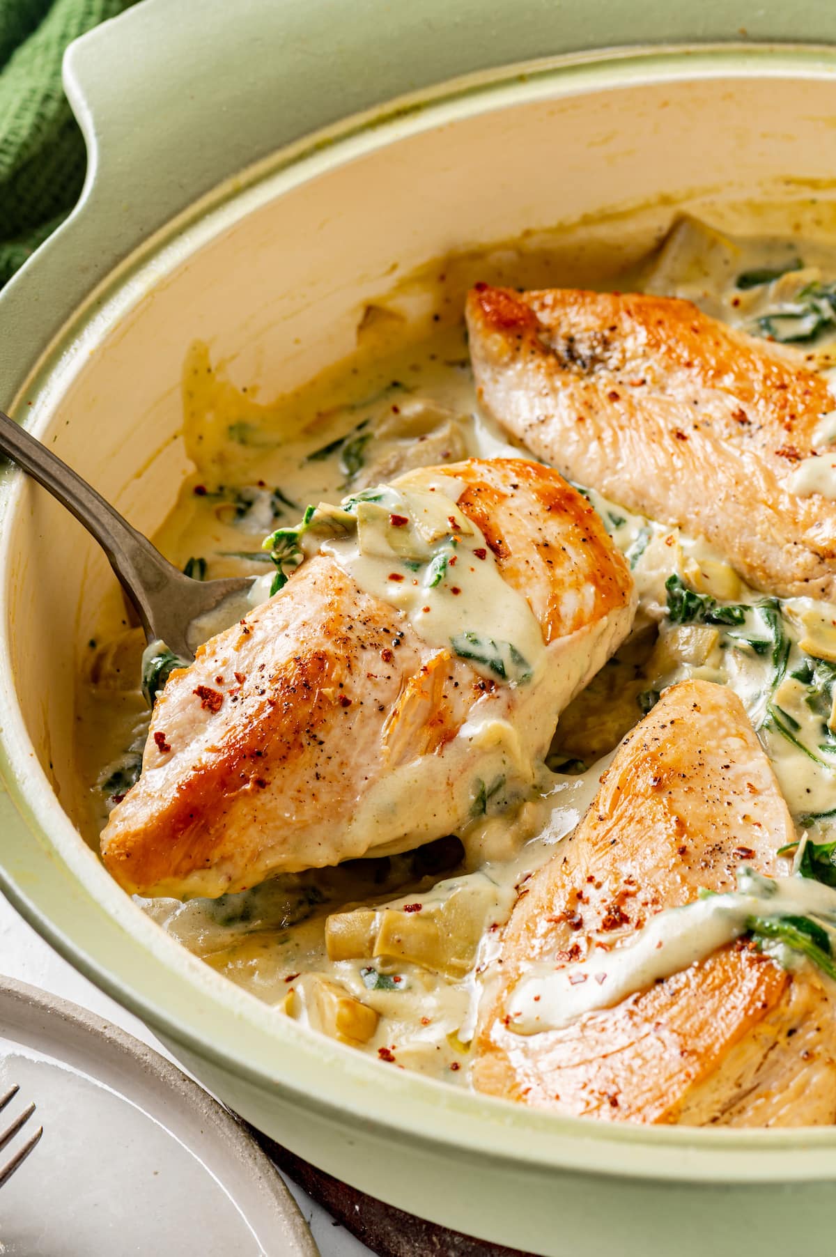 A spoon lifting a saucy chicken piece from creamy spinach artichoke chicken in a large skillet.