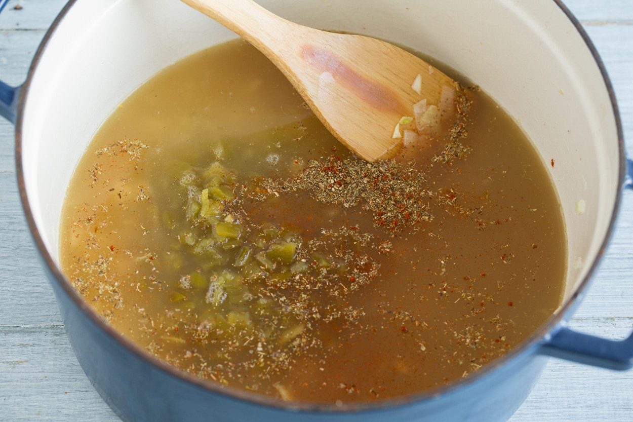 Adding broth, spices and green chilis to white chicken chili mixture in pot.